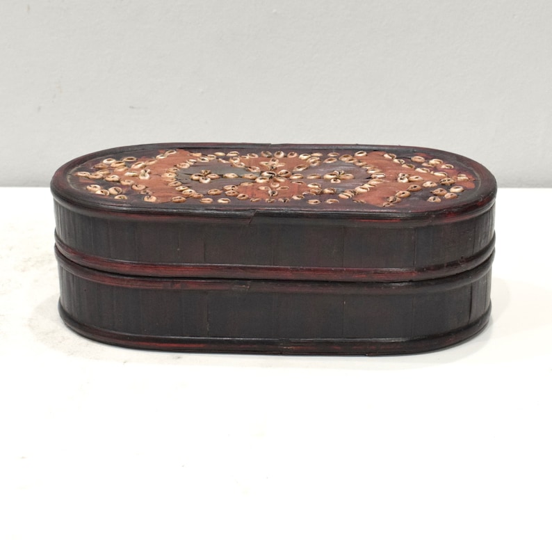 May include: An oval-shaped wooden box with a dark brown finish. The top of the box is decorated with a pattern of small, white shells.