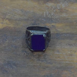 May include: A silver-toned ring with a rectangular, deep blue stone. The ring's band features an openwork design. The ring is set against a wooden surface.