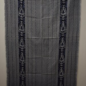 Op de afbeelding: Een grijs en wit geweven deken met zwarte en blauwe geometrische patronen langs de randen. De deken heeft een franje rand.