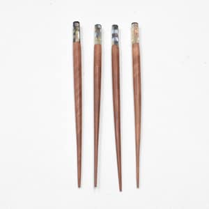 May include: Four wooden hair sticks with decorative tops. The sticks are a warm brown color, and the tops have a mosaic-like design with iridescent accents. The sticks are arranged vertically against a white background.