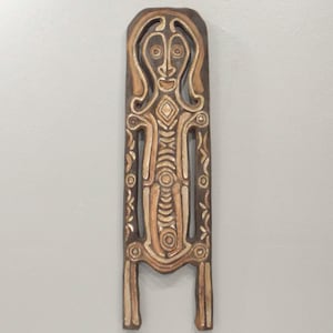 Pode incluir: Uma peça de parede de madeira esculpida com uma figura humana estilizada. A obra de arte é principalmente marrom e bege, com padrões intrincados e um design simétrico. A figura tem uma cabeça arredondada, corpo alongado e duas pernas.