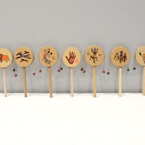 May include: Eight small wooden hand drums with painted designs. The drums have a handle and a small bead attached to the bottom. The designs include a buffalo, a handprint, a bird, and a skull.