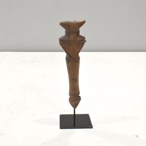 May include: A wooden figurine of a person with a triangular head and a long, slender body. The figurine is standing on a black stand.