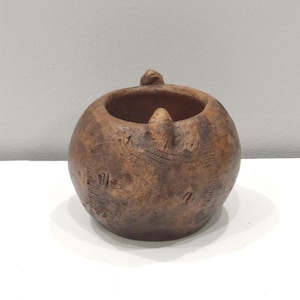 Puede incluir: Una pequeña maceta de cerámica marrón con forma redonda y superficie texturizada. La maceta tiene tres pequeñas figuras en relieve en el borde, y la superficie está decorada con un patrón de líneas y puntos.