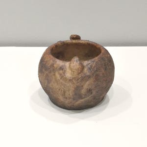 May include: A small, round, brown ceramic pot with a textured surface. The pot has a small opening at the top and a raised lip around the opening.
