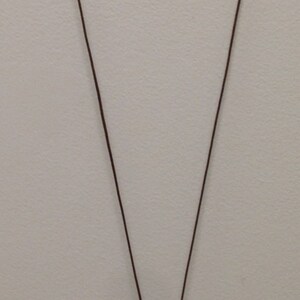 May include: A brown cord necklace with a pendant featuring orange beads, a dark brown geometric bead, and a gold-colored circular charm. The necklace is displayed against a neutral background.