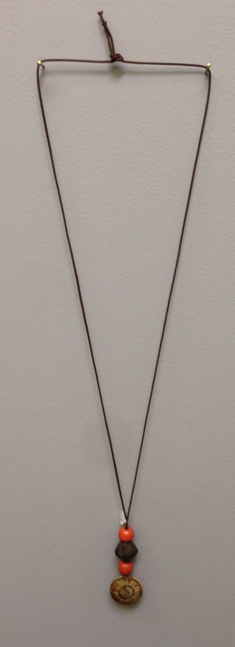 May include: A brown cord necklace with a pendant featuring orange beads, a dark brown geometric bead, and a gold-colored circular charm. The necklace is displayed against a neutral background.