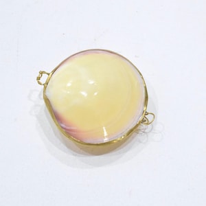 May include: A circular clam shell container with a smooth, polished surface featuring cream, yellow, and soft pink hues. The shell is encased in a thin, gold-toned metal frame with small, delicate wire loops attached to opposite sides of the rim.