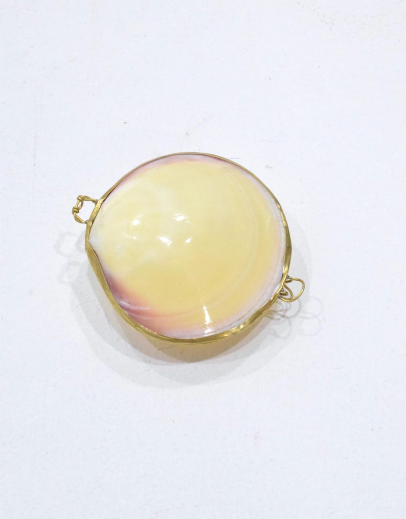 May include: A circular clam shell container with a smooth, polished surface featuring cream, yellow, and soft pink hues. The shell is encased in a thin, gold-toned metal frame with small, delicate wire loops attached to opposite sides of the rim.