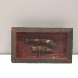 Puede incluir: Un marco de madera con un paspartú de terciopelo rojo y dos cuchillos de aspecto antiguo expuestos en su interior. Los cuchillos tienen mangos de madera y hojas de metal.