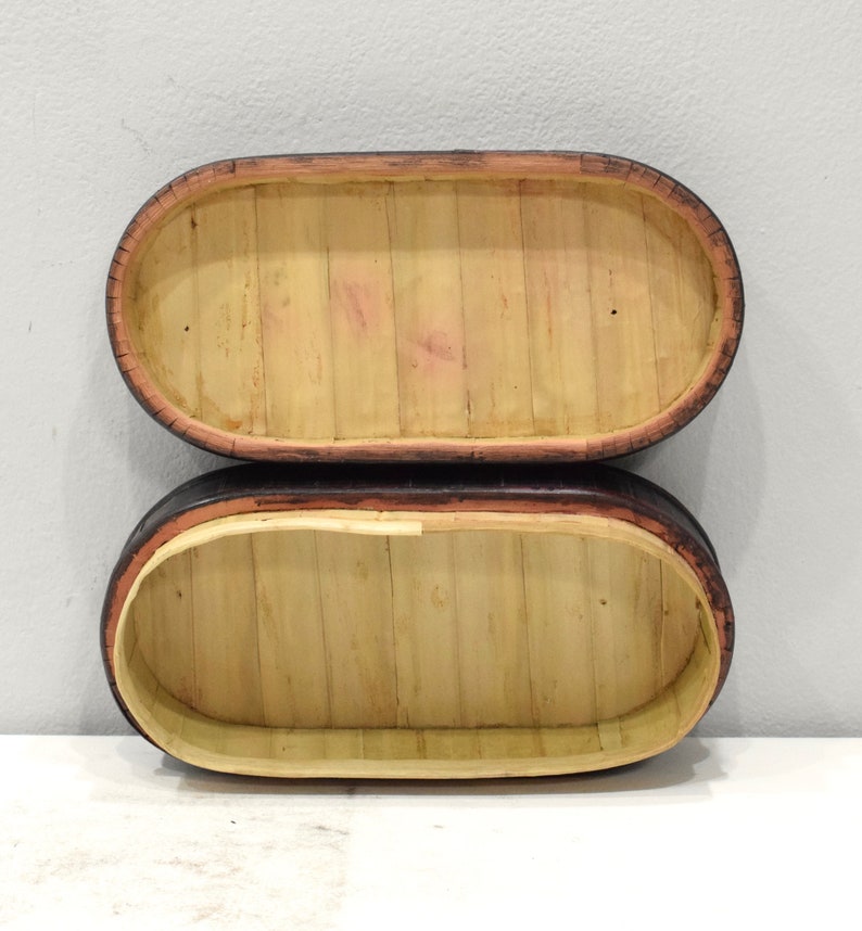 May include: Two oval-shaped, wooden boxes with black and brown trim. The boxes are made of woven bamboo and are stacked on top of each other.