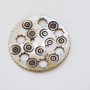 May include: A round, white, mother-of-pearl pendant with a decorative design of concentric circles and holes.