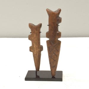 May include: Two wooden carvings of stylized figures on a black stand. The figures are carved in a simple, geometric style and have a brown color.