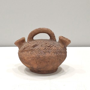 Puede incluir: Una pequeña jarra de cerámica marrón con dos picos y un asa. La jarra tiene una superficie texturizada y está decorada con un patrón tejido.