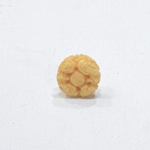 May include: A small, round, ivory-colored bead with intricate carvings. The bead has a floral design with a central motif.