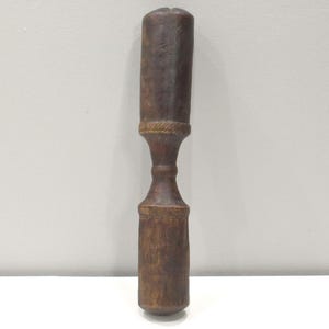 Può includere: Un mazzuolo di legno con una finitura marrone e una forma leggermente a clessidra. Il mazzuolo ha una testa rotonda e liscia e un manico leggermente affusolato.