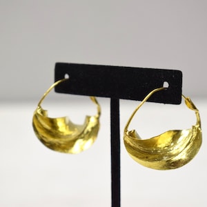 May include: A pair of gold hoop earrings with a wavy, textured design. The earrings are displayed on a black stand.