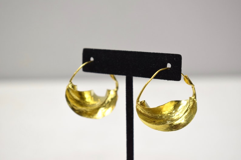 May include: A pair of gold hoop earrings with a wavy, textured design. The earrings are displayed on a black stand.