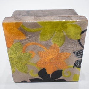 Puede incluir: Una caja cuadrada de cerámica con un diseño floral. La caja tiene un fondo gris con flores naranjas, amarillas y verdes. Las flores tienen un diseño texturizado y en relieve.