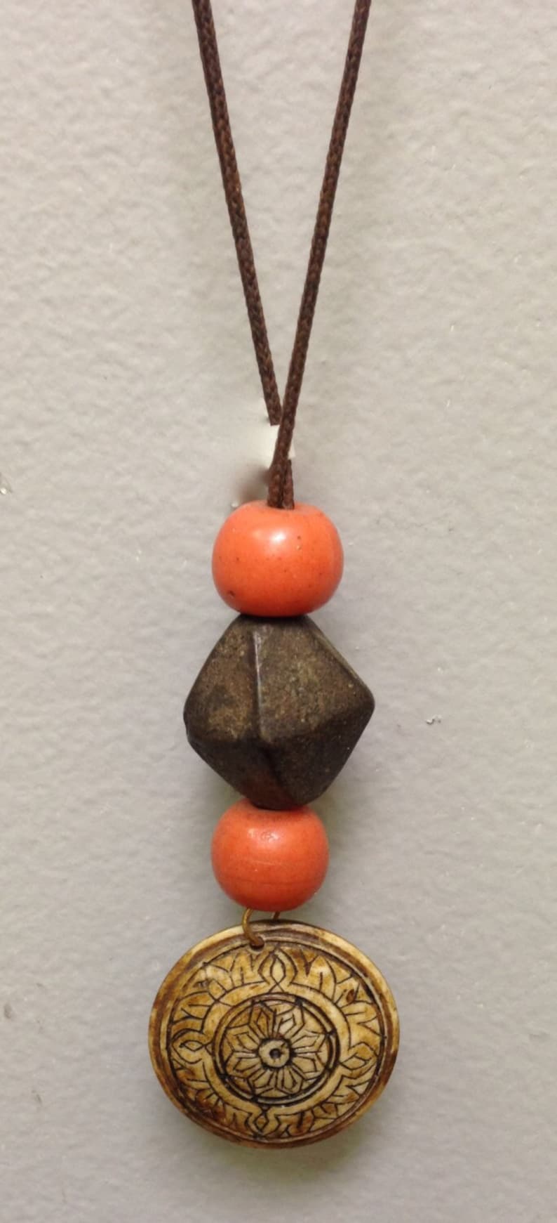 May include: A handcrafted necklace featuring a brown cord, orange beads, a dark geometric bead, and a carved circular pendant with an intricate floral design. The pendant is a light tan color.