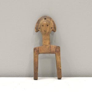 Puede incluir: Una figurita de madera de una persona con una cara estilizada y un cuerpo simple y geométrico. La figurita está tallada en madera marrón claro y tiene un agujero en la parte superior de la cabeza.