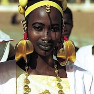 May include: A woman wearing traditional African jewelry, including a large gold necklace, earrings, and a headpiece with yellow beads. She has a black and white design painted on her face.