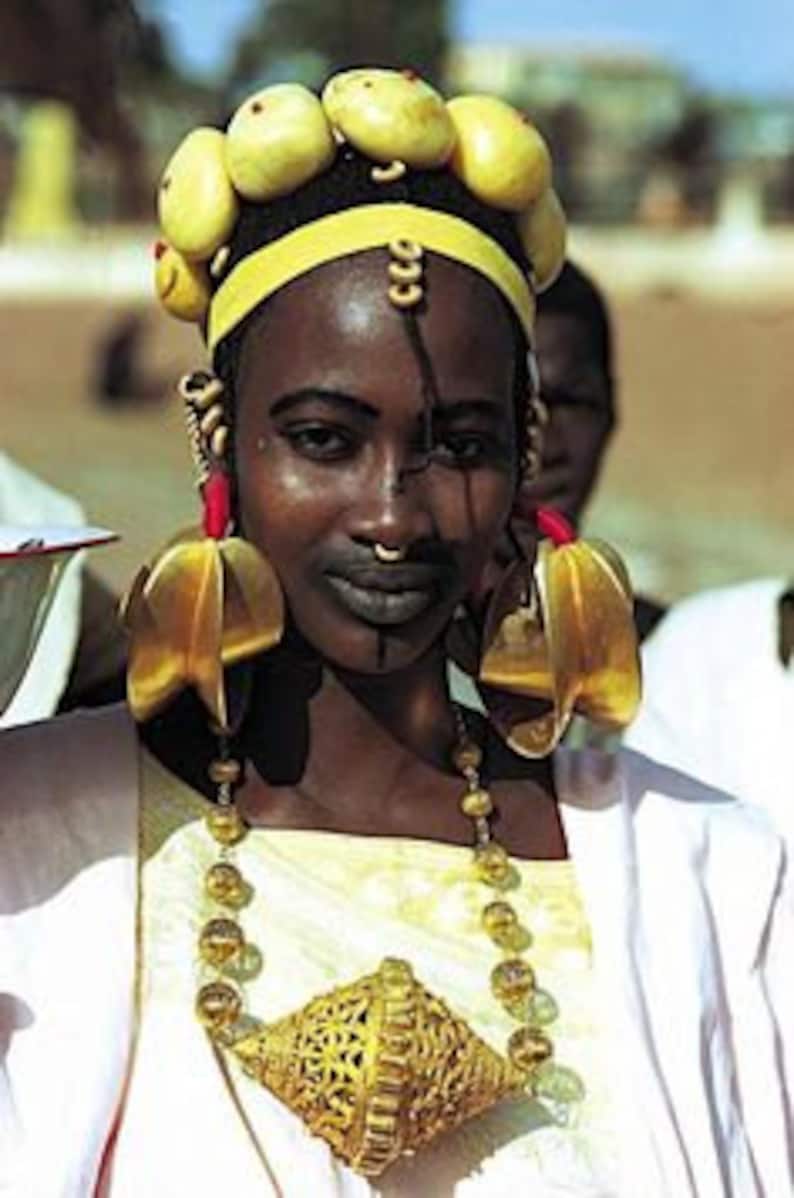 May include: A woman wearing traditional African jewelry, including a large gold necklace, earrings, and a headpiece with yellow beads. She has a black and white design painted on her face.
