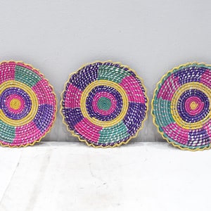 Könnte beinhalten: Drei runde geflochtene Teller mit einem farbenfrohen Design. Die Teller sind aus natürlichen Materialien gefertigt und haben ein kreisförmiges Muster in Pink, Lila, Grün und Gelb.