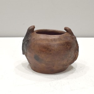 Puede incluir: Una pequeña maceta de cerámica marrón con dos asas en forma de cabezas de animales estilizadas. La maceta tiene una superficie rugosa y texturizada y parece estar hecha a mano.