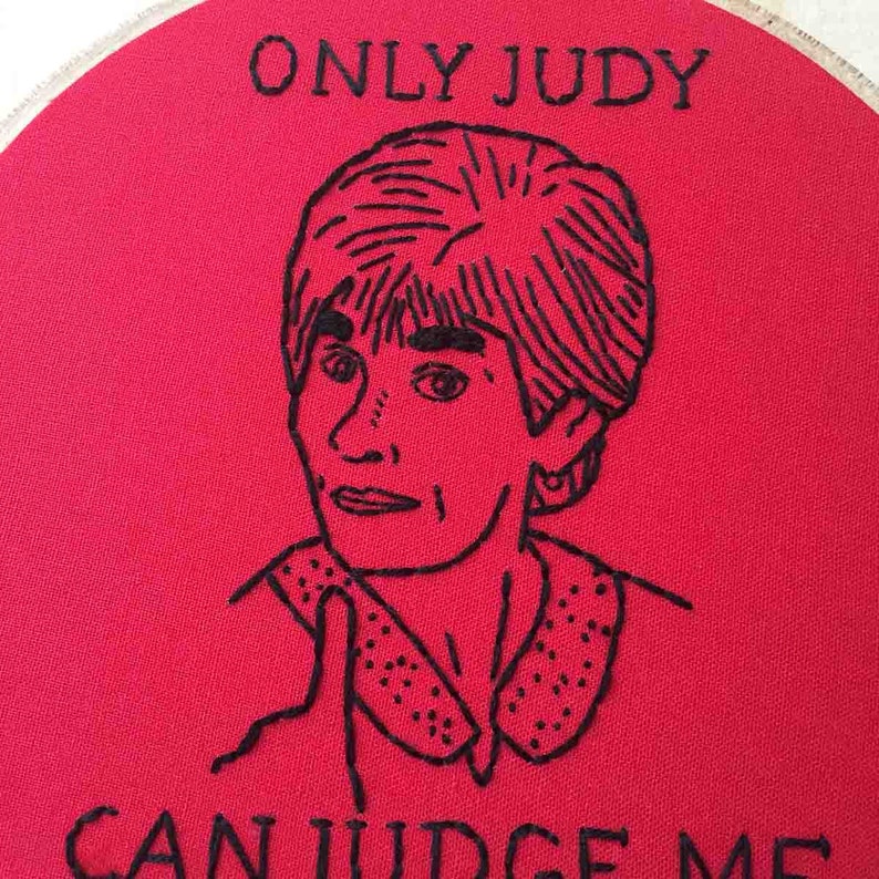 May include: Embroidered hoop art with a red background and black embroidery. The embroidery depicts a person with short hair and the text "ONLY JUDY CAN JUDGE ME".