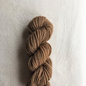 Puede incluir: Una madeja de hilo marrón, bien enrollada y torcida, sobre una superficie blanca. El hilo parece ser de fibra natural, con un aspecto ligeramente texturizado. Ideal para tejer, hacer ganchillo u otros proyectos de arte textil.