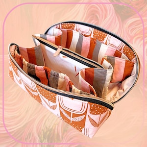 May include: Open, semi-circular fabric organizer with multiple compartments. The exterior features a white background with brown floral and dot patterns. Interior compartments have striped and solid fabric in shades of brown, gray, and pink. A gold zipper secures the organizer.