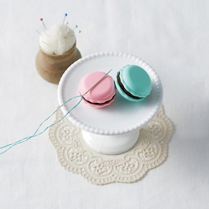 May include: Two macarons, one pink and one blue, are displayed on a white cake stand. A needle and thread are threaded through the pink macaron.