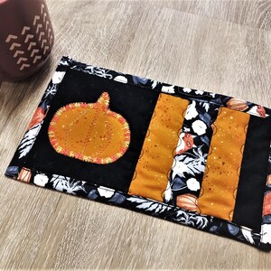 May include: A rectangular, quilted coaster with a pumpkin applique. The coaster features a black background with orange and floral accents. An orange pumpkin with yellow stitching is centered on the coaster. A maroon mug is in the background.