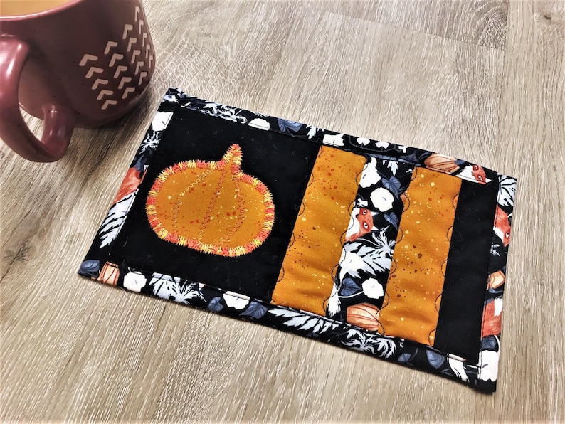 May include: A rectangular, quilted coaster with a pumpkin applique. The coaster features a black background with orange and floral accents. An orange pumpkin with yellow stitching is centered on the coaster. A maroon mug is in the background.