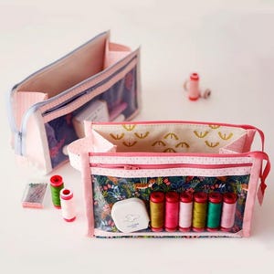 May include: Two pink and navy blue sewing pouches with clear panels, one open and filled with spools of thread, a white Clover measuring tape, and needles. The other pouch is closed. The pouches have a floral pattern.