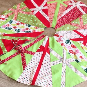 May include: A round Christmas tree skirt with a festive design. The skirt features a patchwork of red, green, and white fabrics with various patterns, including polka dots and geometric shapes. Red ribbon ties secure the skirt.