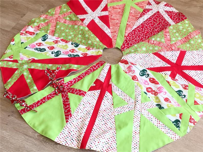 May include: A round Christmas tree skirt with a festive design. The skirt features a patchwork of red, green, and white fabrics with various patterns, including polka dots and geometric shapes. Red ribbon ties secure the skirt.