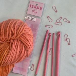 May include: A collection of crochet supplies, including a skein of peach-colored yarn, pink crochet hooks, and pink stitch markers. The packaging reads "LYKKE Crafts" and "Blush Wooden Hooks". The crochet hooks are US M13, 9.00mm.