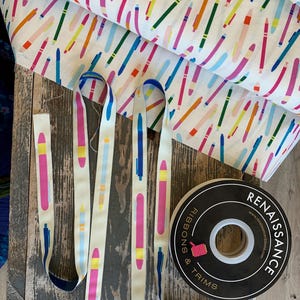 Op de afbeelding: Een verzameling knutselbenodigdheden met stof en decoratieve linten met een kleurrijk pennenontwerp op een witte achtergrond. Een zwart-witte rol lint is ook zichtbaar. De items liggen op een houten oppervlak.