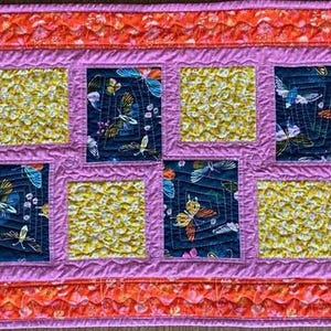May include: A quilted table runner with a repeating pattern of squares. The squares alternate between a dark blue fabric with butterfly designs and a yellow, textured fabric. The runner has a pink border and an orange outer edge.