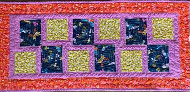 May include: A quilted table runner with a repeating pattern of squares. The squares alternate between a dark blue fabric with butterfly designs and a yellow, textured fabric. The runner has a pink border and an orange outer edge.