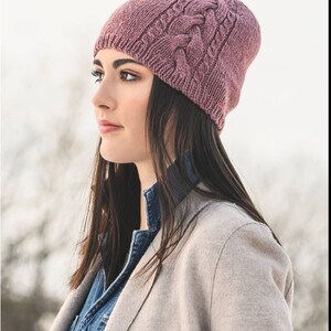 May include: A woman wearing a mauve knitted beanie with a cable knit pattern. The beanie is pulled down over her ears and she is looking off to the side.
