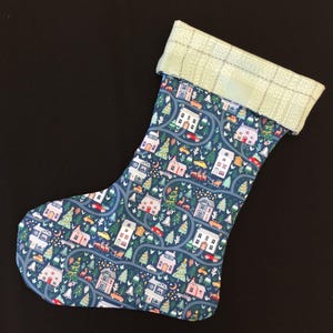 May include: A Christmas stocking with a blue background featuring a repeating pattern of houses, trees, and cars. The cuff is a light green plaid. The stocking is made of fabric.