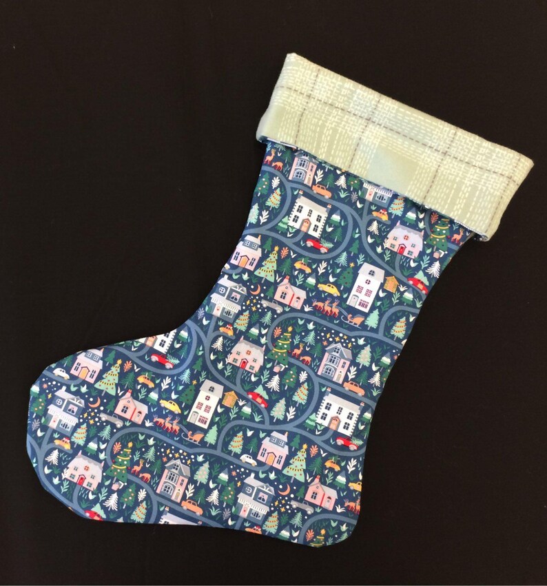 May include: A Christmas stocking with a blue background featuring a repeating pattern of houses, trees, and cars. The cuff is a light green plaid. The stocking is made of fabric.