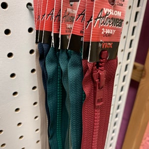 Può includere: Primo piano di una varietà di tiretti per cerniere lampo in diversi colori, tra cui rosso, verde e blu. Le cerniere sono etichettate "VISLON Activewear 2-Way Cherry" e "By YKK".