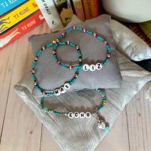 Könnte beinhalten: Drei Perlenarmbänder mit türkisfarbenen, braunen und rosafarbenen Perlen. Die Armbänder haben die Namen "Max", "Liz" und "Echo" mit weißen Buchstabenperlen ausgeschrieben. Das "Echo"-Armband hat einen kleinen Herzanhänger.