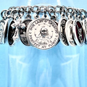 May include: A silver charm bracelet with multiple religious charms, including a cross, a depiction of Jesus, and a charm that reads "Our Lady of Loreto I Protect My Flight".