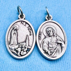 May include: Silver oval medal with two sides. One side features an image of the Virgin Mary with the text "N.S. Fatima". The other side features an image of Jesus with his heart exposed.