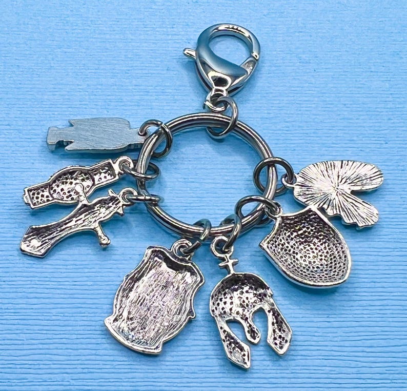 May include: Silver keychain with multiple charms, including a shield, a helmet, a lily pad, a cross, and a barrel. The charms are attached to a silver ring with a silver lobster clasp.